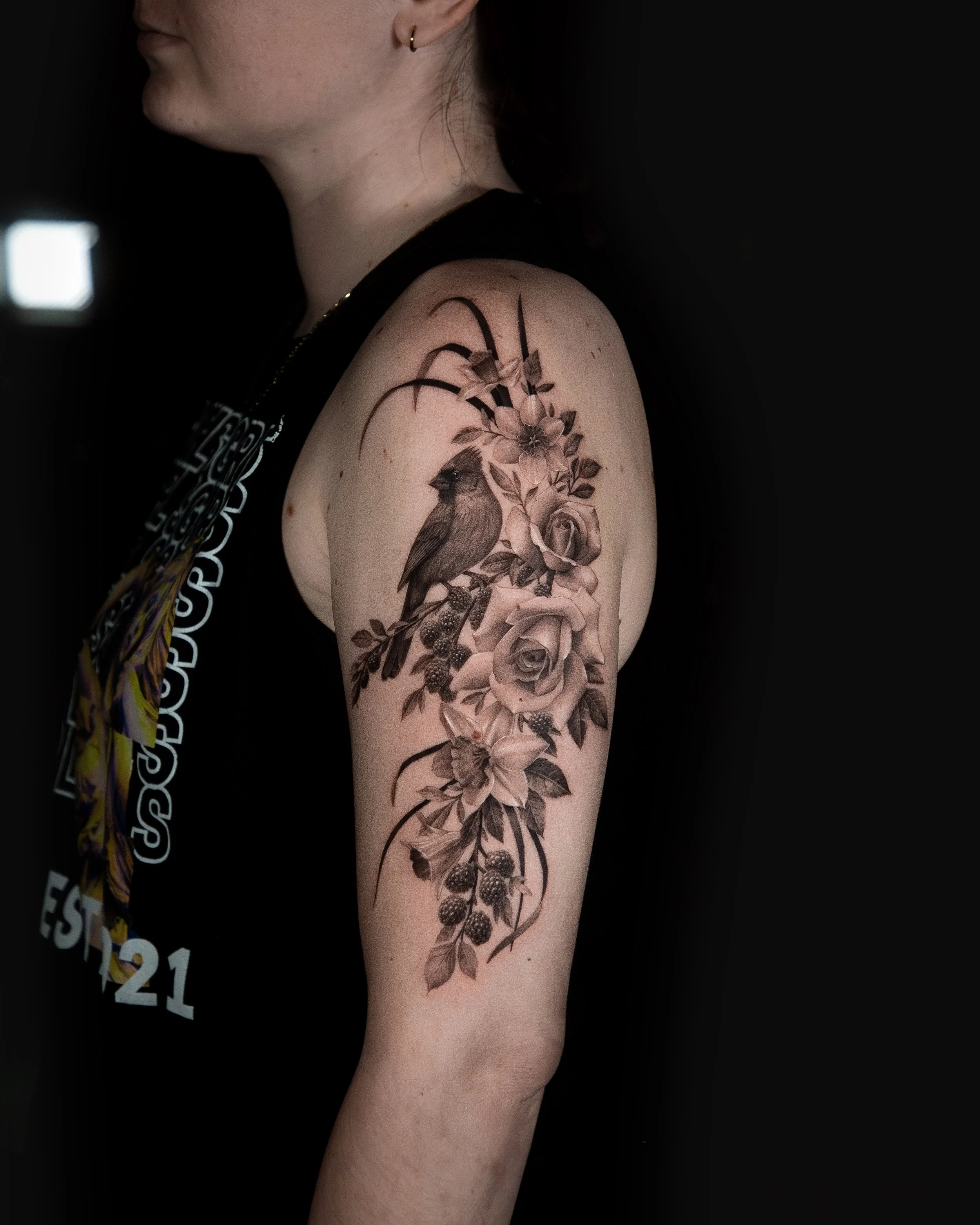 A hyper realistic tattoo done by Ihor (flowerhead.ink) on a person's shoulder. Featuring a close-up of a person's upper arm showing a detailed black-and-grey tattoo of a bird, flowers, leaves, and berries.