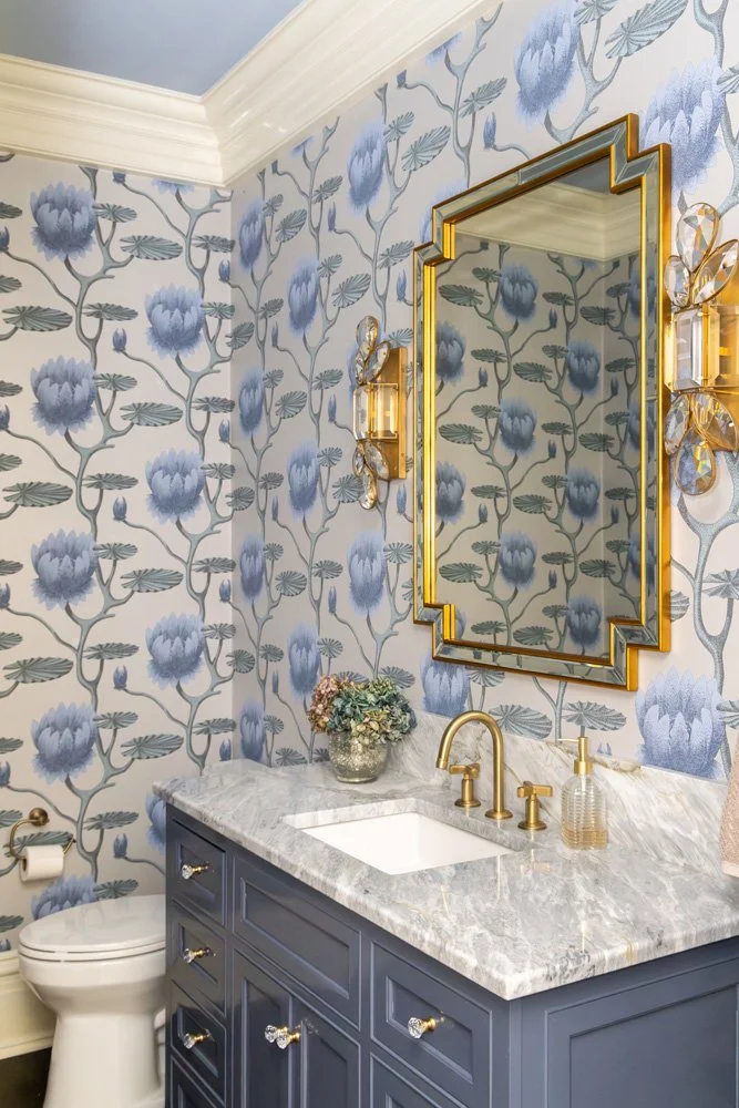Elegant powder room with blue floral wallpaper, navy vanity, marble countertop, gold fixtures, and geometric framed mirror