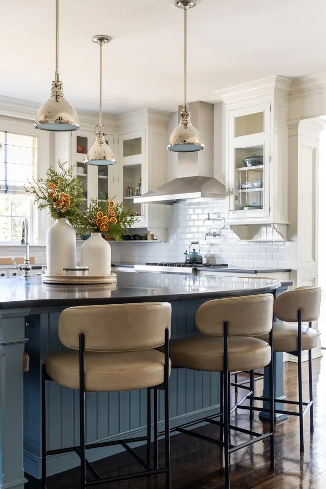 Bright traditional kitchen with navy island, white cabinets, pendant lights, marble countertops, and upholstered bar stools
