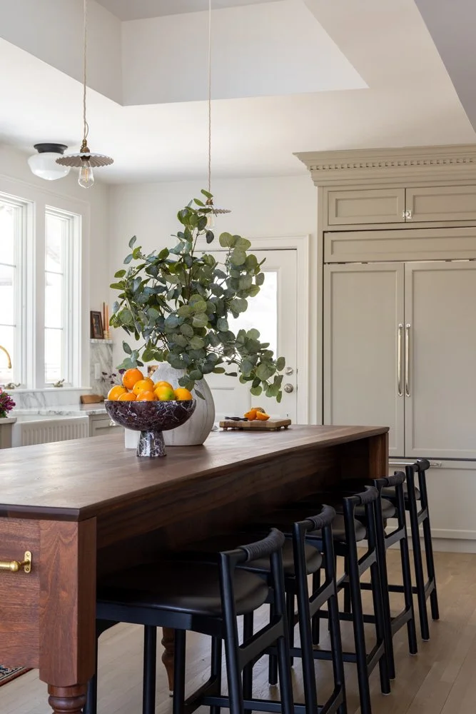 Bright modern kitchen with a dark wood island, black bar stools, gray cabinetry, pendant lights, and a bowl of oranges with leafy branches.