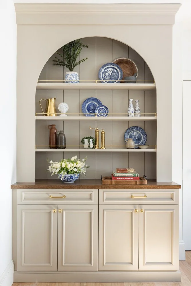 Cream built-in hutch with arched shelving styled with blue-and-white ceramics, brass accents, flowers, and books