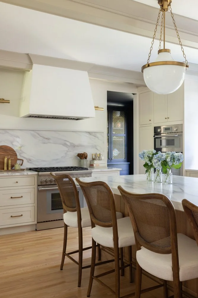 Elegant white kitchen with marble island, cane bar stools, pendant light, and blue hydrangea centerpiece