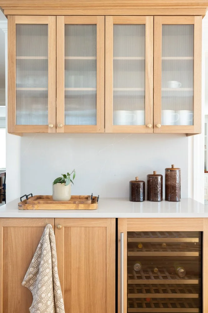 Bright kitchen with oak cabinets, glass-front upper cupboards, white countertop, potted plant, ceramic jars, and built-in wine cooler