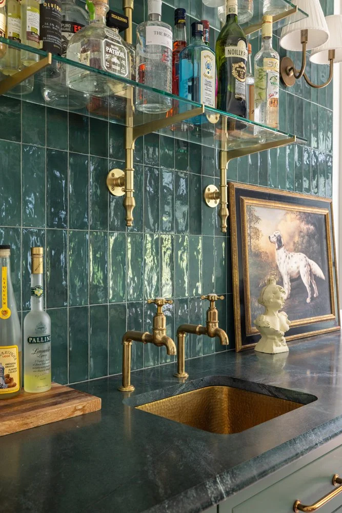 Elegant home bar with liquor bottles on glass shelves, brass faucets, dark green tile backsplash, farmhouse sink, framed dog painting, and white statuette