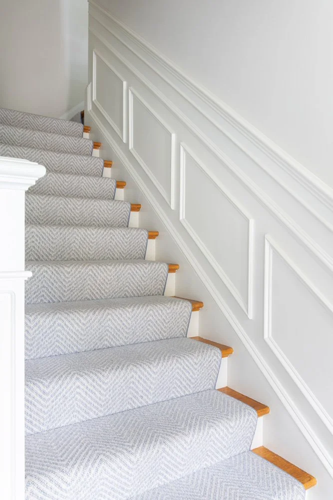 Light gray patterned carpet runner on a white staircase with natural wood stair edges and decorative wall paneling