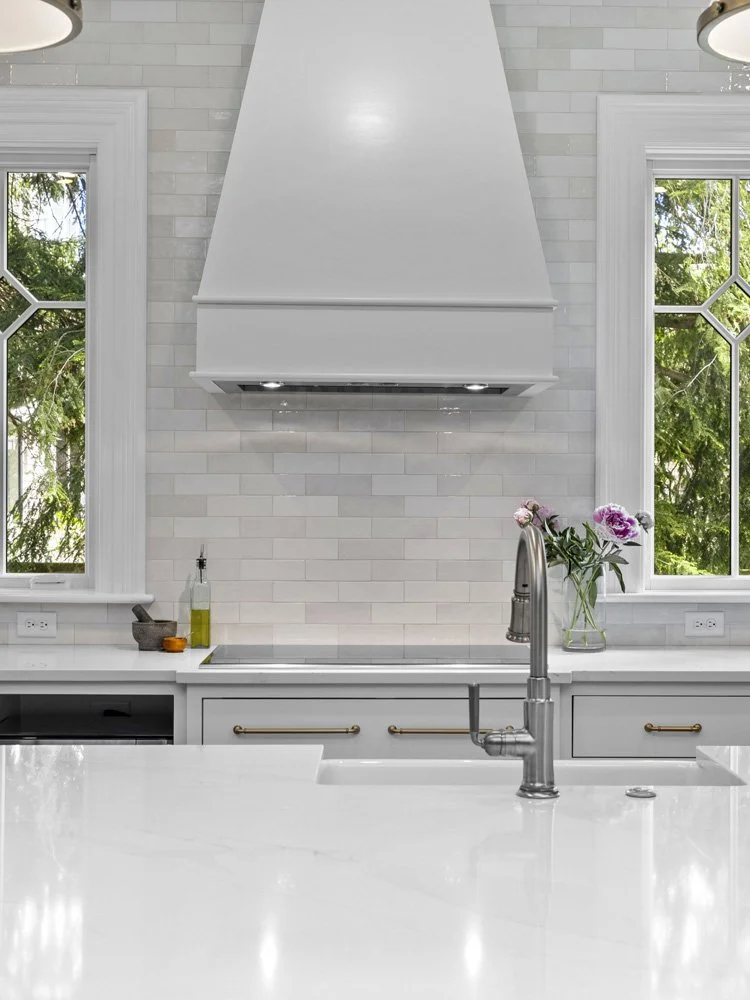Bright white kitchen with marble island, farmhouse sink, brass cabinet hardware, subway tile backsplash, and large range hood