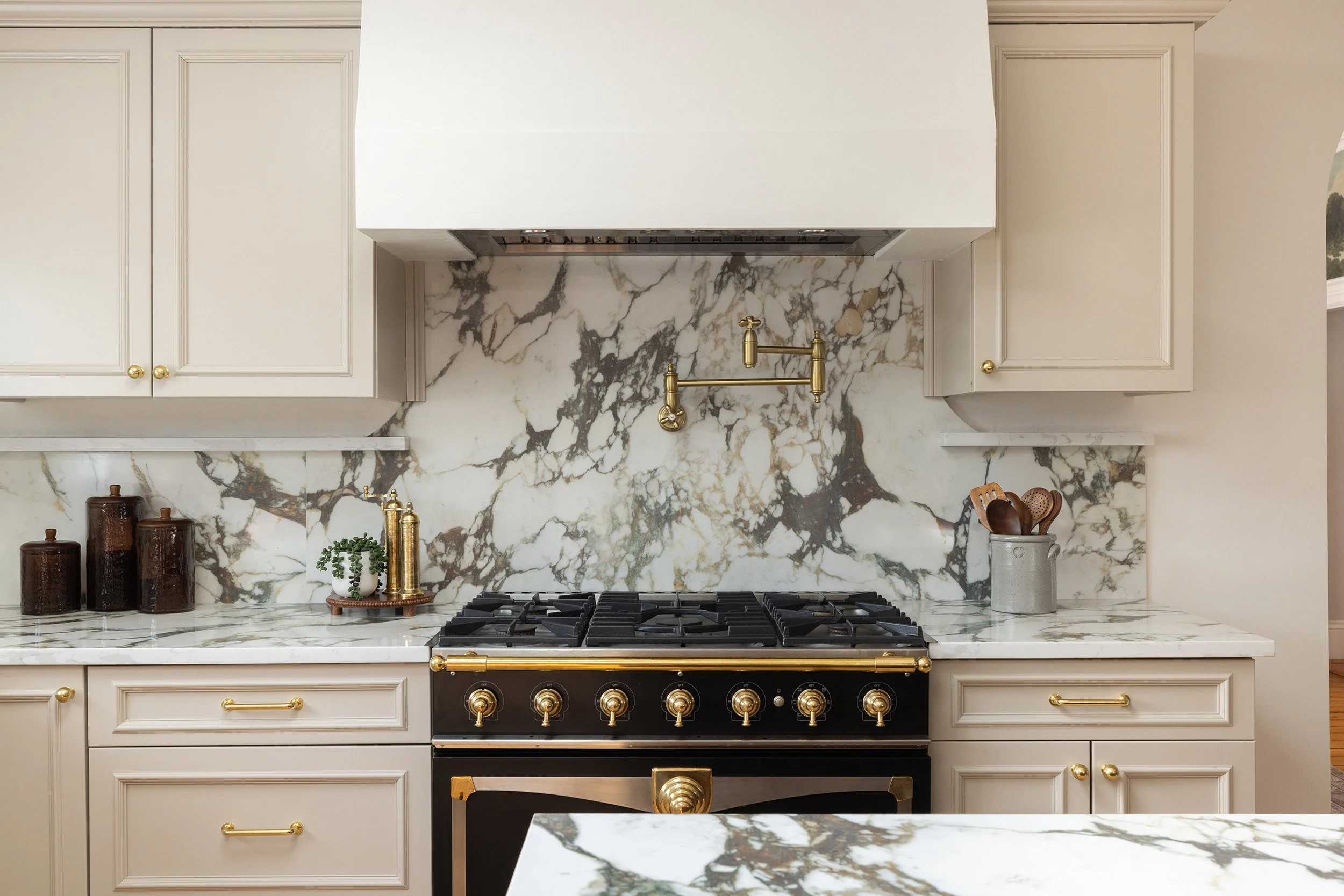 Elegant cream kitchen with marble backsplash and countertops, black and brass gas range, hood, gold fixtures, and wooden utensils.