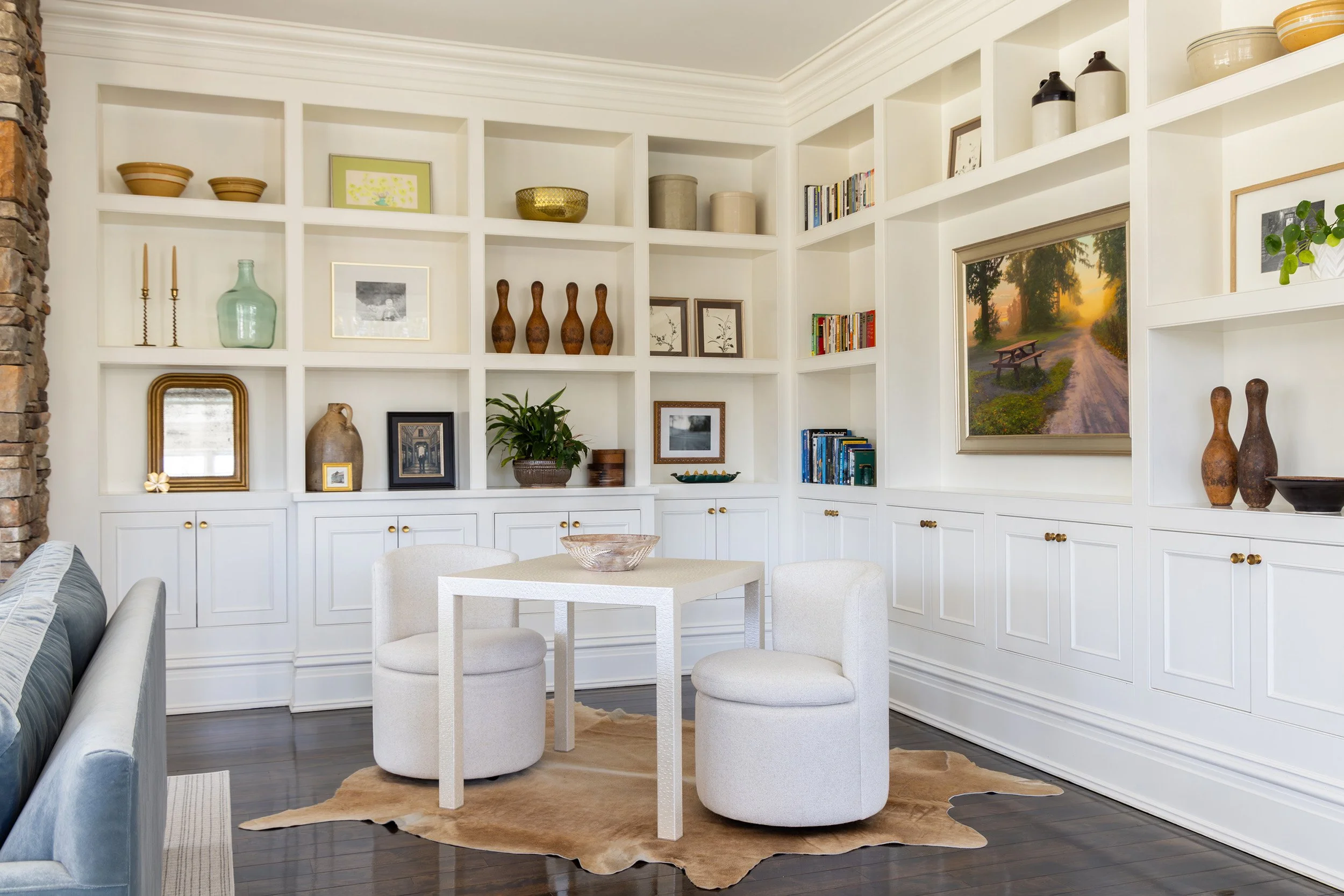 Bright living room with built-in white display shelves, artwork, books, decorative pottery, a painting, and two white chairs around a square table.