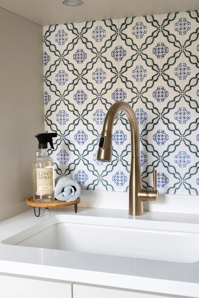 White bathroom sink with a brushed brass gooseneck faucet, patterned blue-and-white tile backsplash, linen spray bottle, and rolled towel