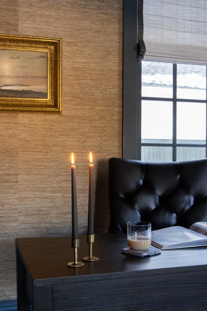 Elegant dark wood desk with two lit taper candles, a glass of coffee, papers, and a black tufted leather chair beside a window