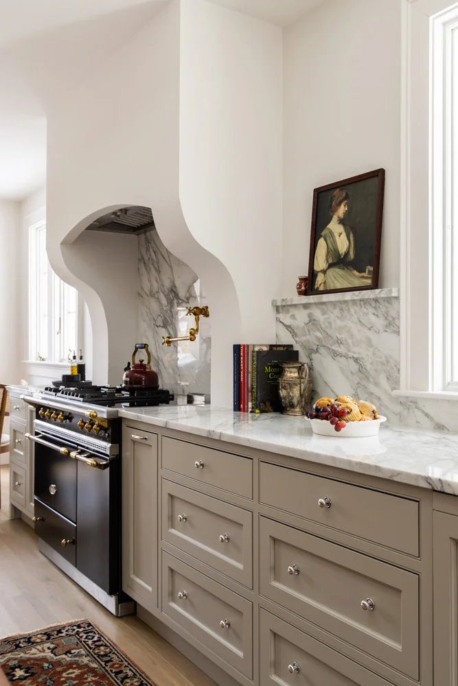 Elegant traditional kitchen with marble backsplash and countertops, black range, taupe cabinetry, fruit bowl, cookbooks, and framed portrait