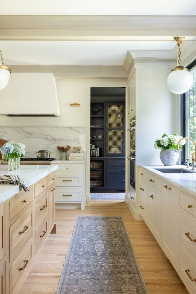 Bright traditional kitchen with white cabinets, marble countertops, central island, floral arrangements, pendant lights, and a patterned runner rug