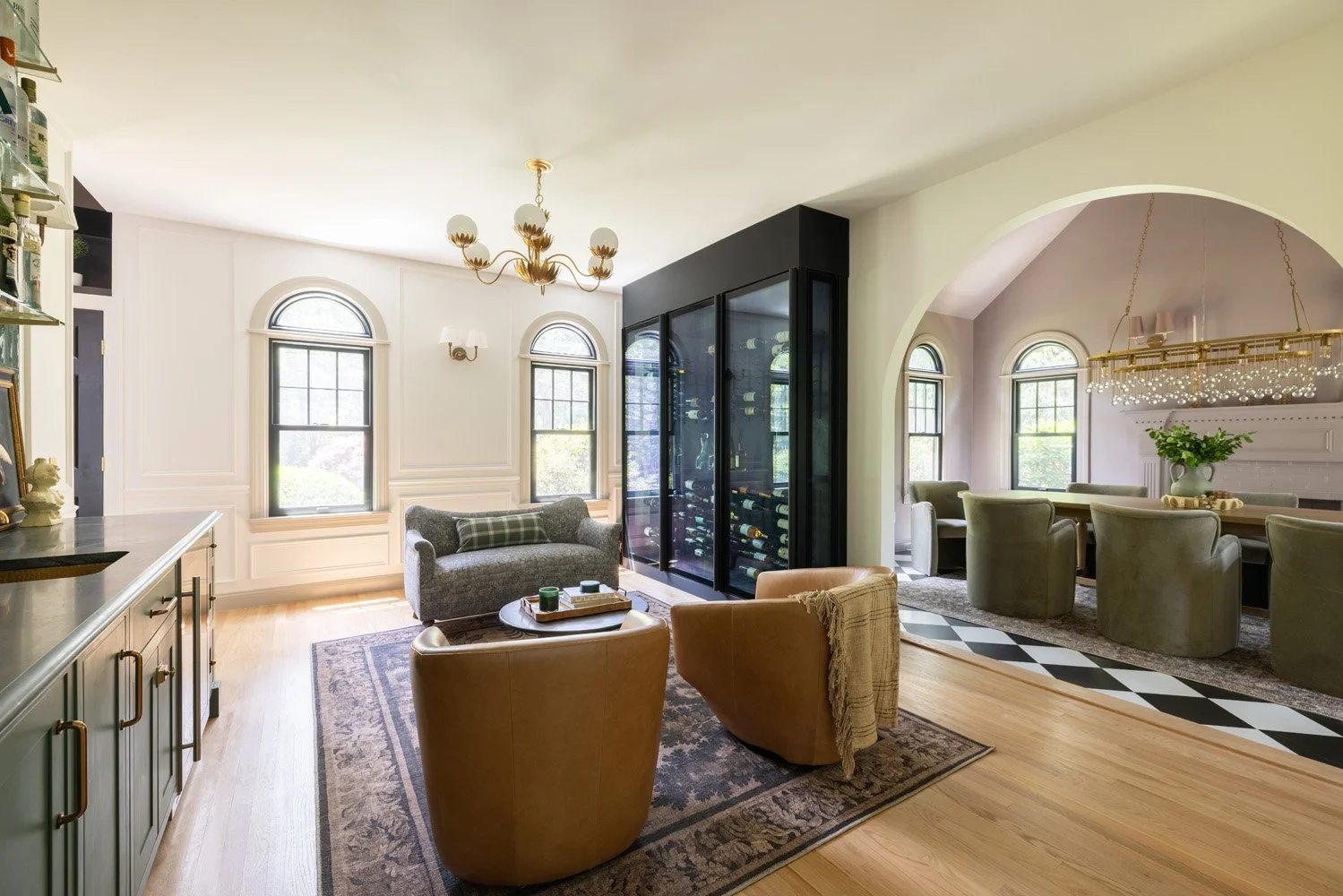 Elegant open-plan living room and dining area with a black glass wine cabinet, leather chairs, arched windows, chandeliers, and hardwood floors