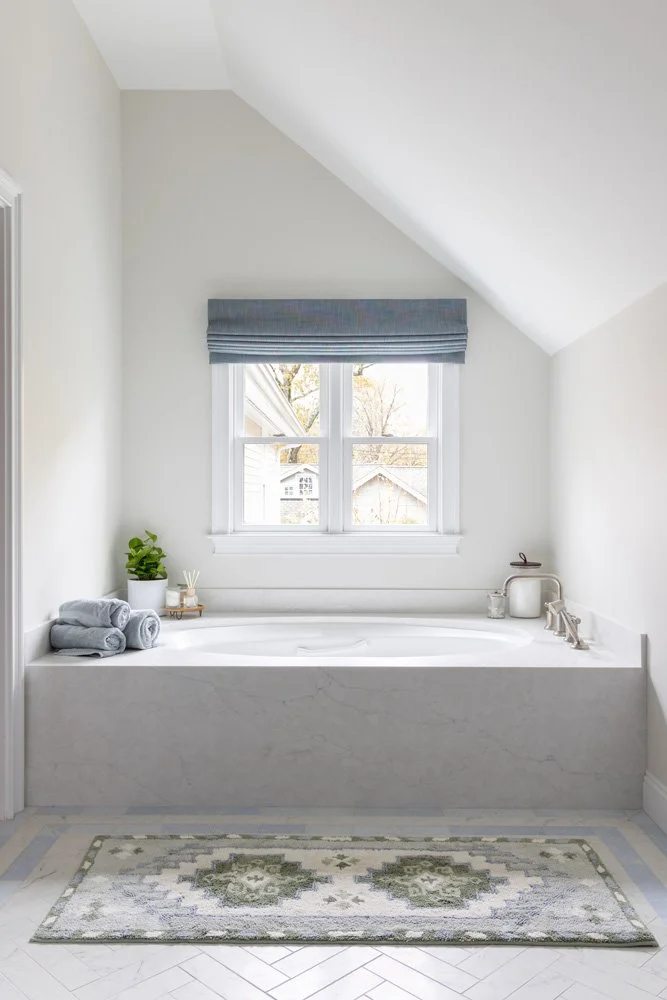 Bright white bathroom with a built-in soaking tub beneath a window, gray Roman shade, folded towels, potted plant, and patterned area rug