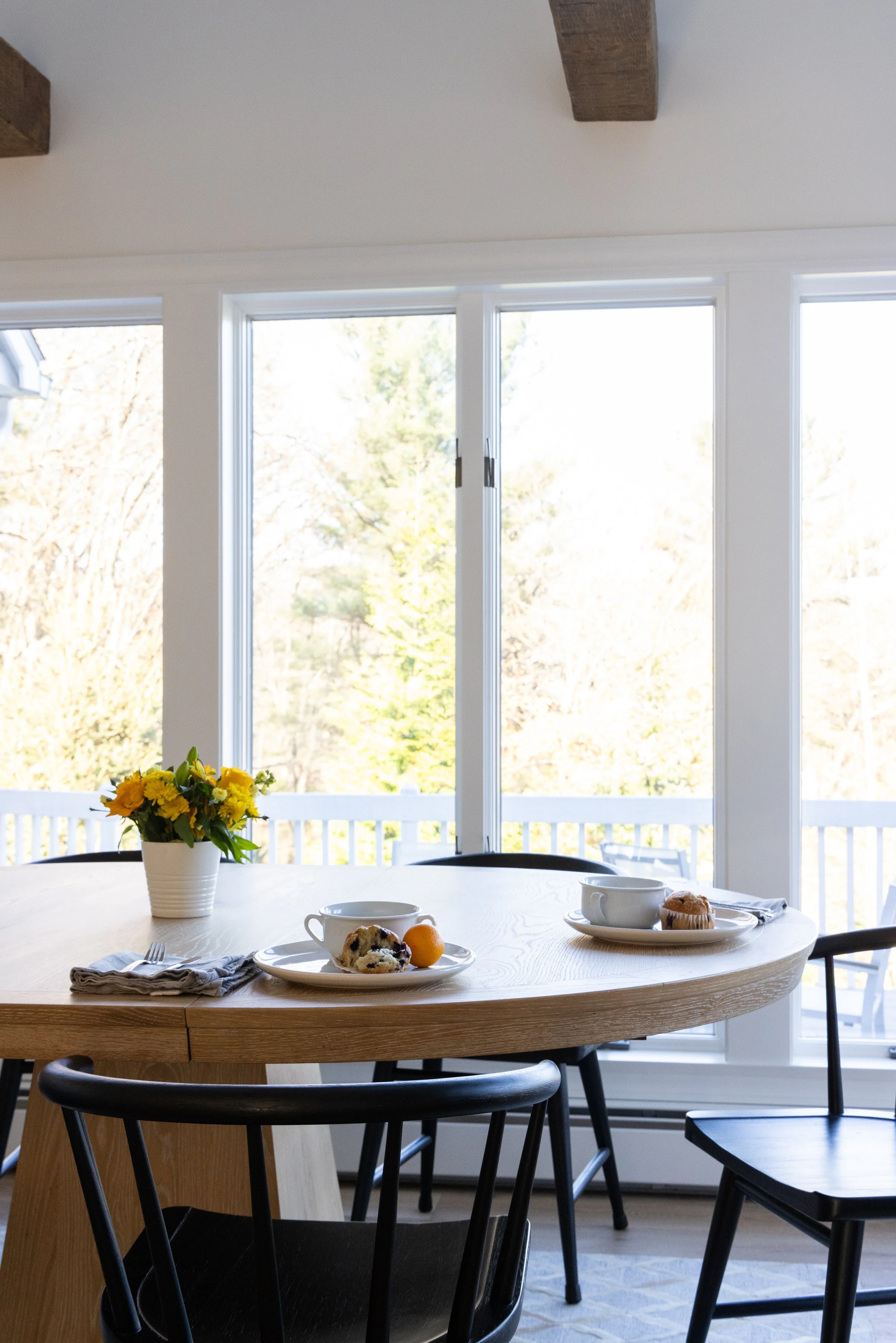 Bright dining room with a wooden table set for breakfast, flowers, coffee cups, muffins, fruit, and large windows overlooking trees.