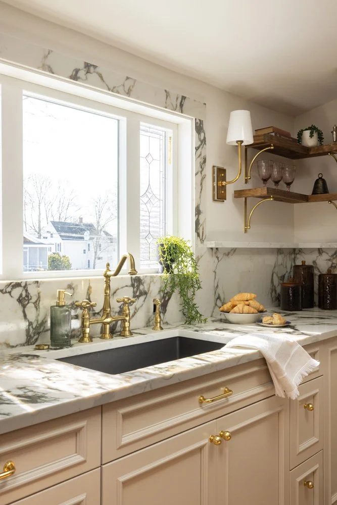 Elegant kitchen with marble countertops and backsplash, farmhouse sink, brass fixtures, open wood shelves, and pastries by a window