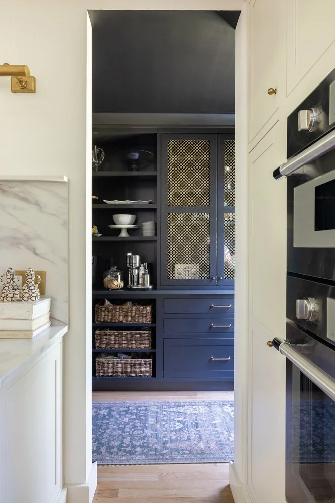 Elegant kitchen butler’s pantry with navy built-in cabinets, brass mesh doors, open shelving, woven baskets, and double wall ovens
