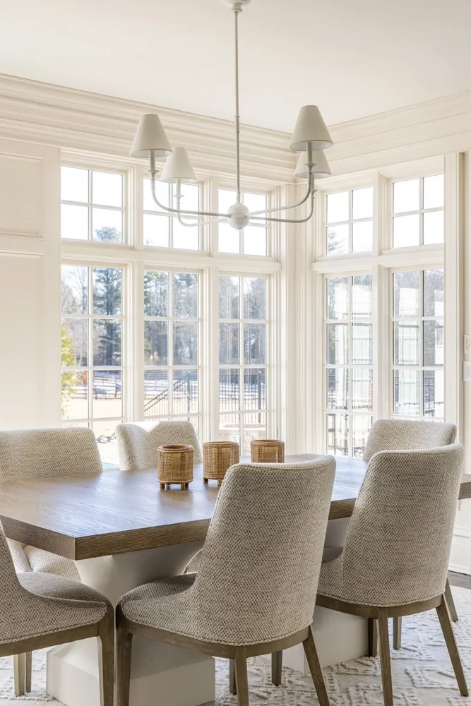 Bright dining room with a wooden table, upholstered chairs, modern chandelier, and large windows.