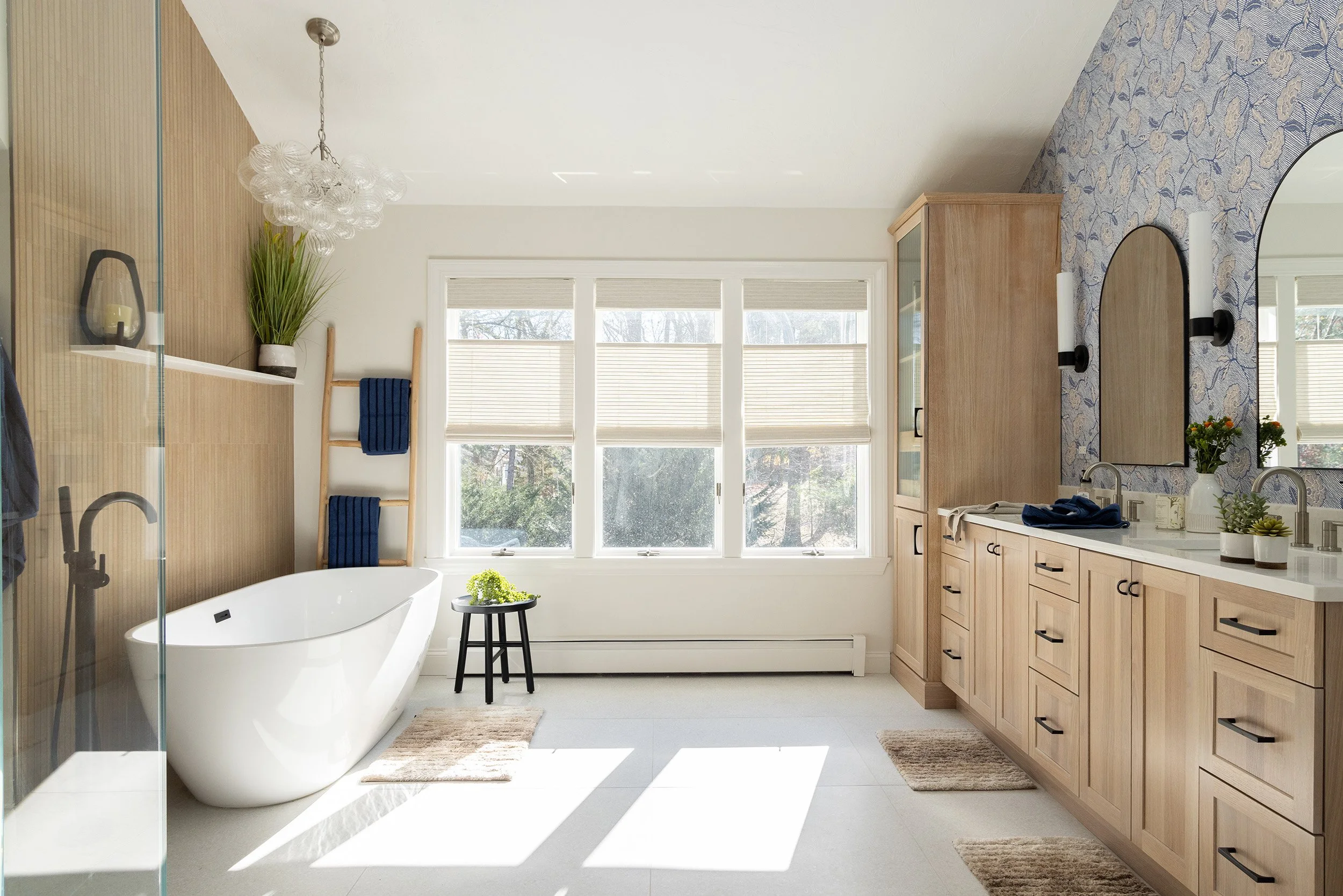 Bright modern bathroom with freestanding bathtub, glass shower, double vanity, floral wallpaper, and large windows.
