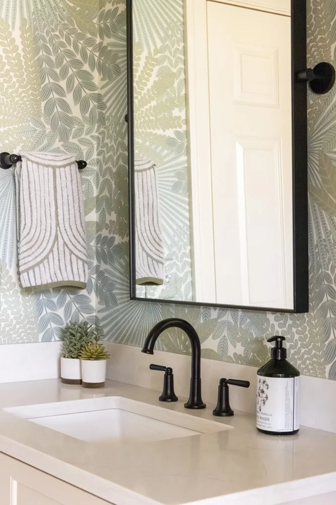 Bathroom vanity with black faucet, framed mirror, patterned botanical wallpaper, striped hand towel, succulents, and soap dispenser