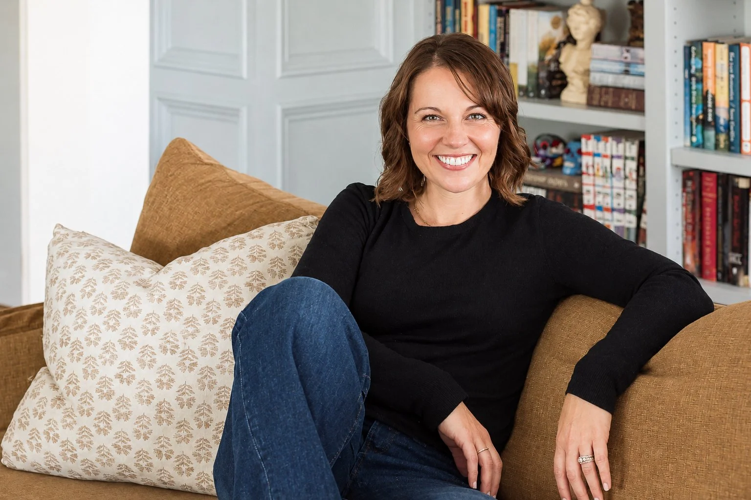 Smiling woman in a black sweater and jeans sitting on a couch with patterned pillows and bookshelves behind her