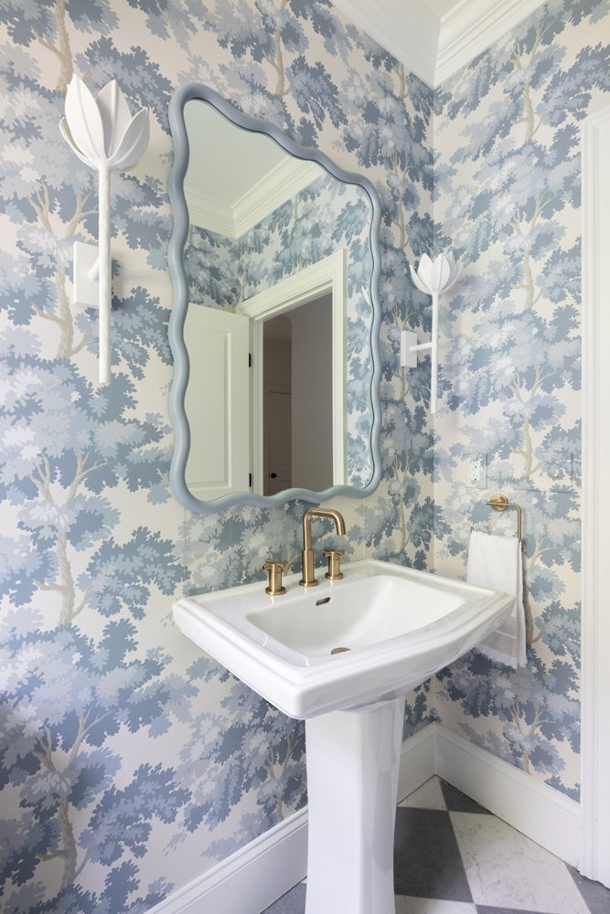 Elegant powder room with blue floral wallpaper, white pedestal sink, wavy-framed mirror, and flower-shaped wall sconces