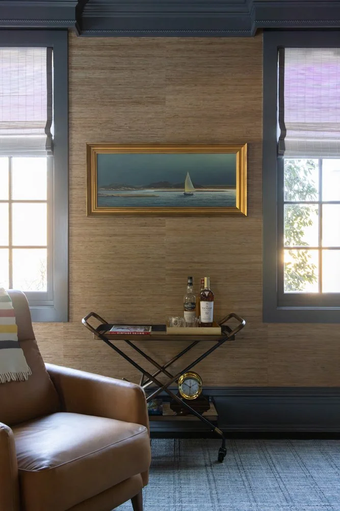 Elegant sitting room with a leather armchair, bar cart with whiskey bottles, sailboat painting, and dark-trimmed windows