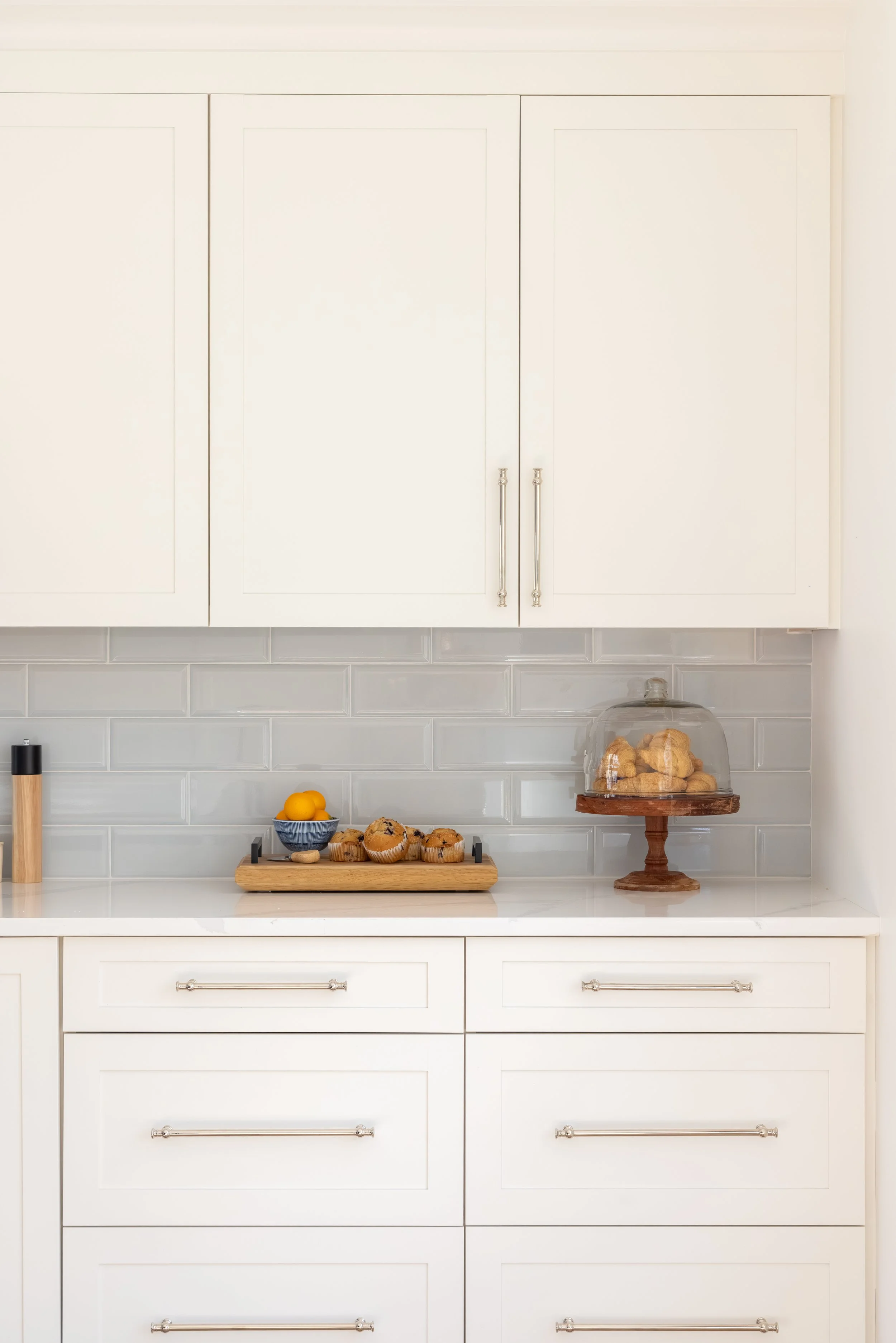 Bright white kitchen with paneled cabinets, subway tile backsplash, muffins, lemons, and pastries displayed on the countertop.