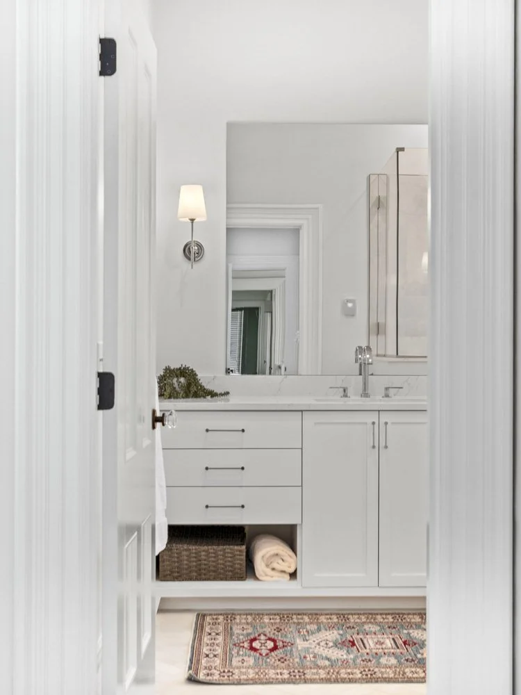 Bright white bathroom vanity with marble countertop, wall mirror, glass shower, cabinets, woven basket, rolled towel, and patterned rug