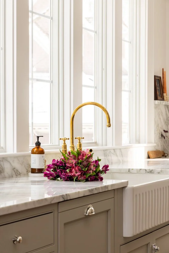 Elegant marble bathroom vanity with gold faucet, pink flower arrangement, soap dispenser, and large windows