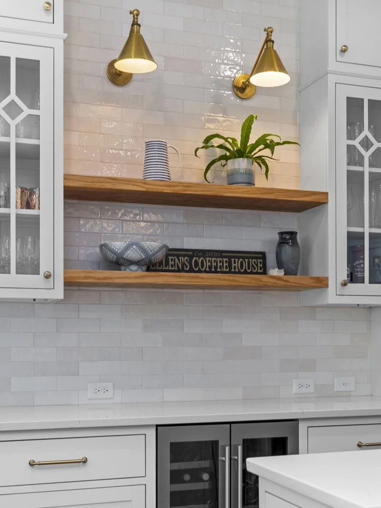 White kitchen with glossy subway tile backsplash, brass sconces, wood shelves, glass cabinets, houseplant, and beverage refrigerator