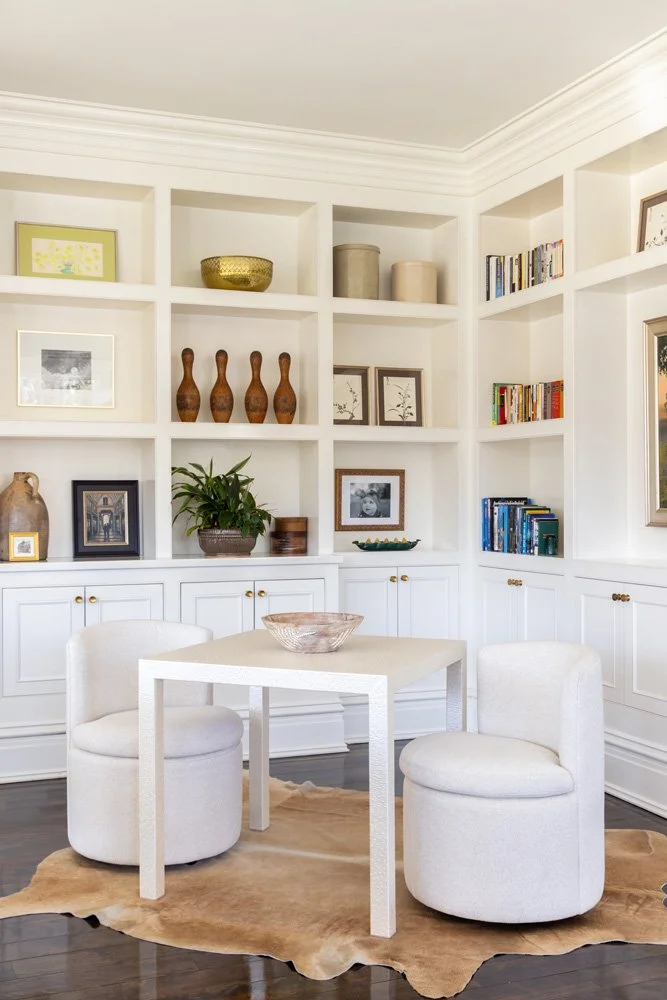 Bright white home office with built-in shelving, books, decorative vases, framed artwork, a small white table, and two boucle chairs on a hide rug