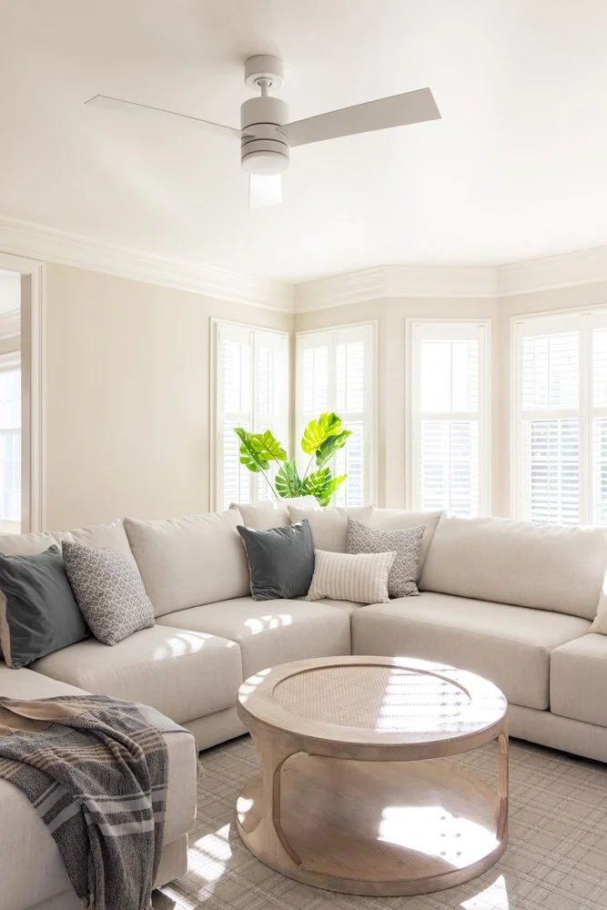 Bright living room with a white sectional sofa, round wooden coffee table, ceiling fan, bay windows, and potted plant