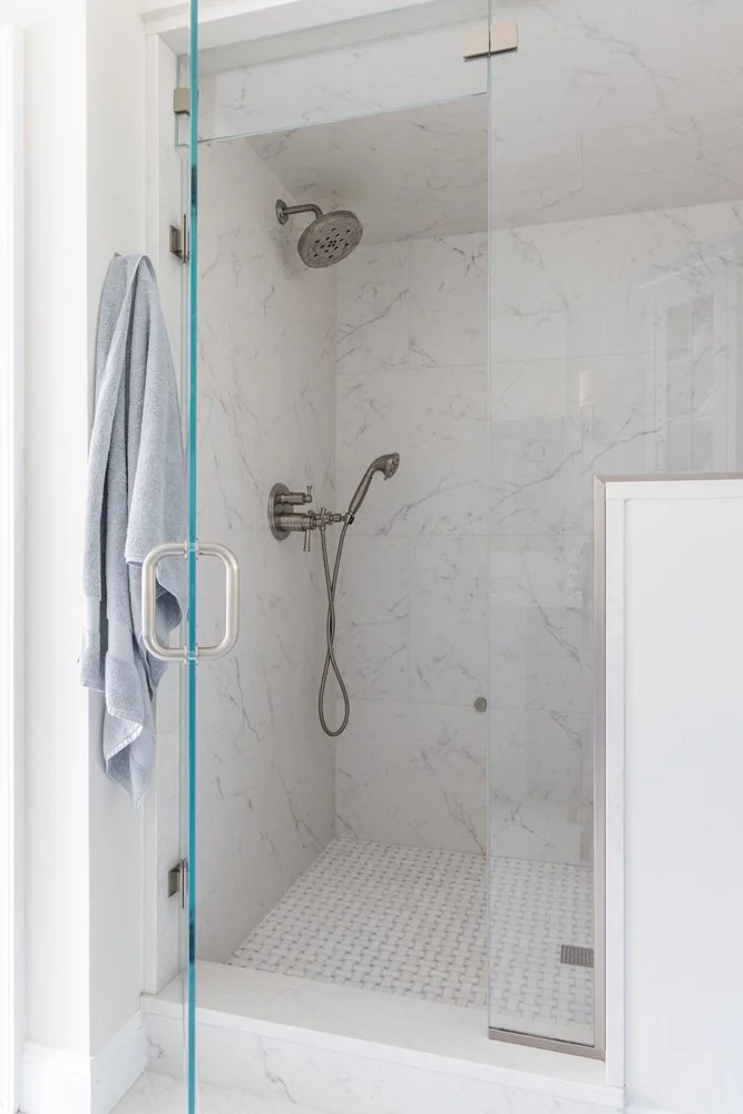 Modern glass-enclosed walk-in shower with white marble tile, handheld and overhead showerheads, and mosaic floor tiles