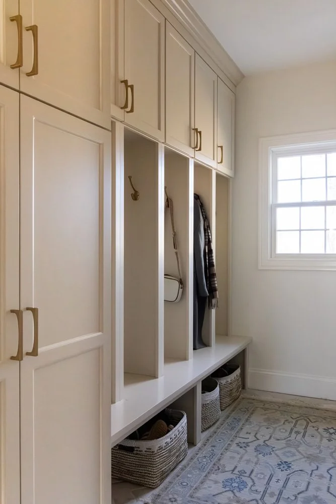 White built-in mudroom cabinets with coat hooks, bench seating, woven storage baskets, and a window