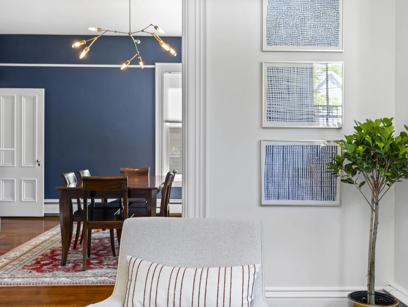 Modern dining room with navy accent wall, wooden table, contemporary chandelier, framed abstract artwork, and potted tree