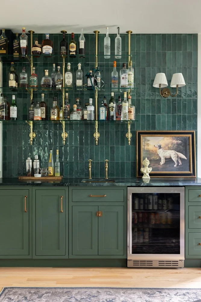 Elegant home bar with green tile backsplash, brass shelves stocked with liquor bottles, framed dog artwork, and built-in beverage refrigerator.