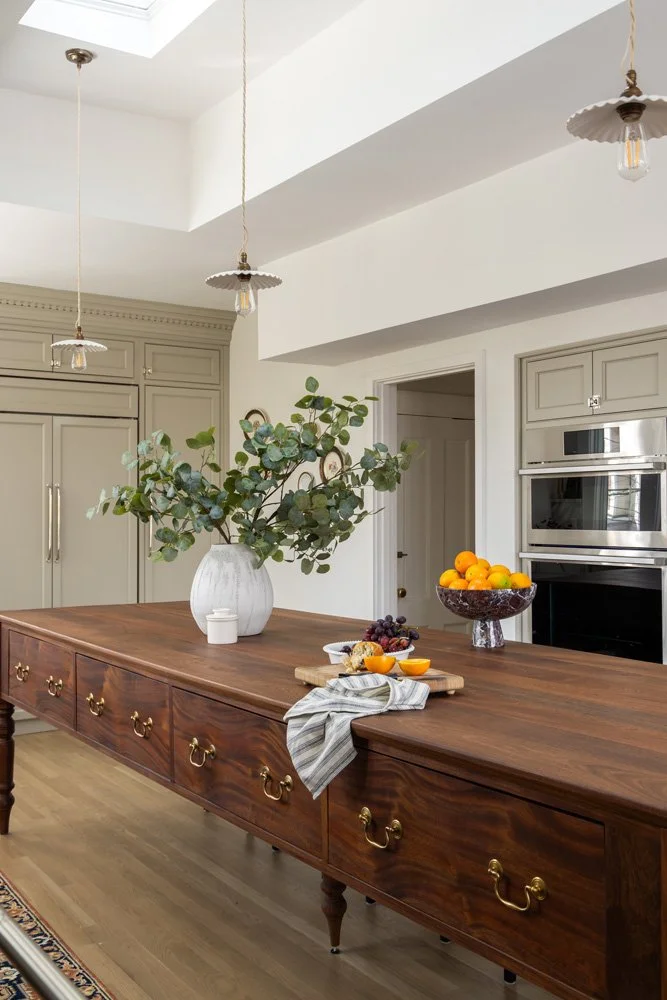 Elegant kitchen with a large wooden island, brass hardware, pendant lights, eucalyptus arrangement, oranges, grapes, and built-in double ovens