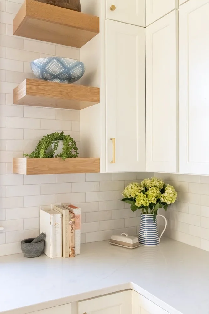 Bright kitchen corner with white subway tile backsplash, floating wood shelves, white cabinets, decorative bowl, potted plant, books, mortar and pestle, and blue-striped pitcher of flowers.