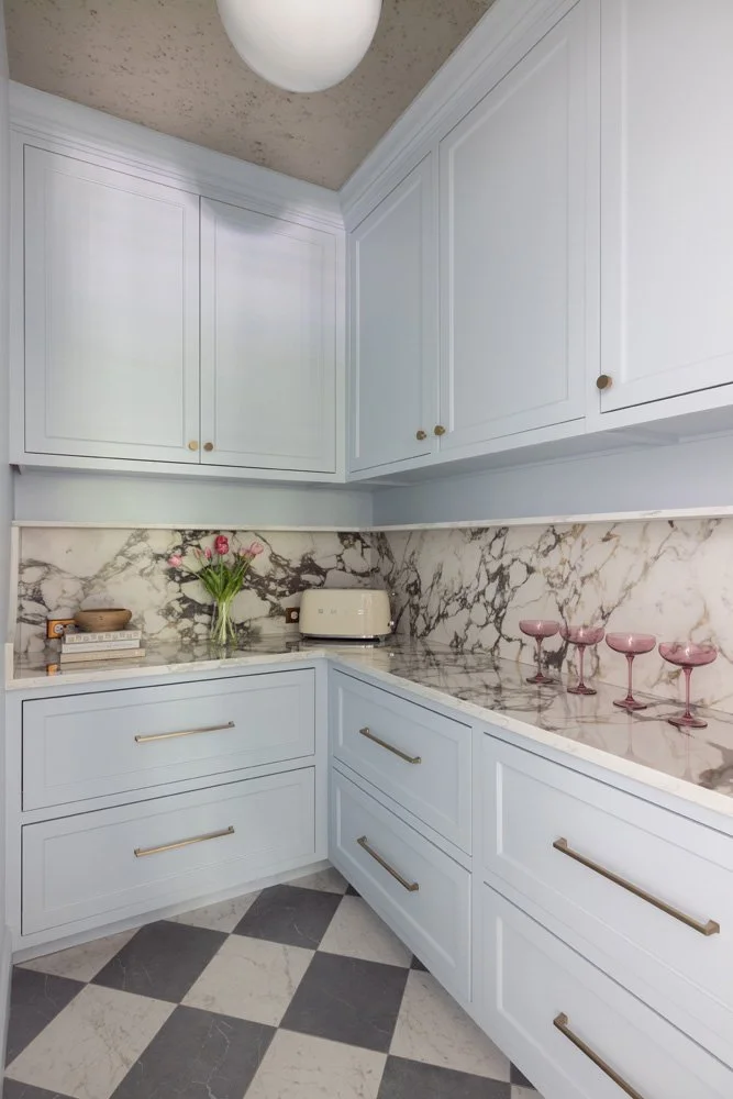 Elegant light blue kitchen with marble backsplash and countertops, brass hardware, toaster, flowers, and pink cocktail glasses