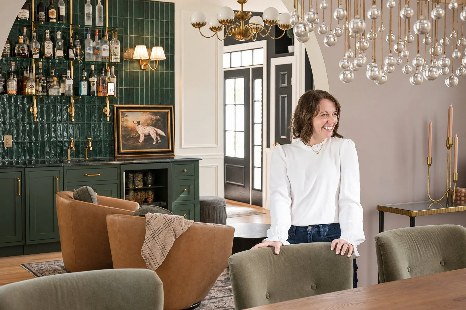 Smiling woman standing in an elegant home bar and dining room with liquor shelves, leather chairs, and modern chandeliers