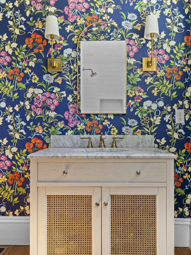 Elegant bathroom vanity with marble countertop, cane-front cabinet doors, gold fixtures, arched mirror, and floral navy wallpaper