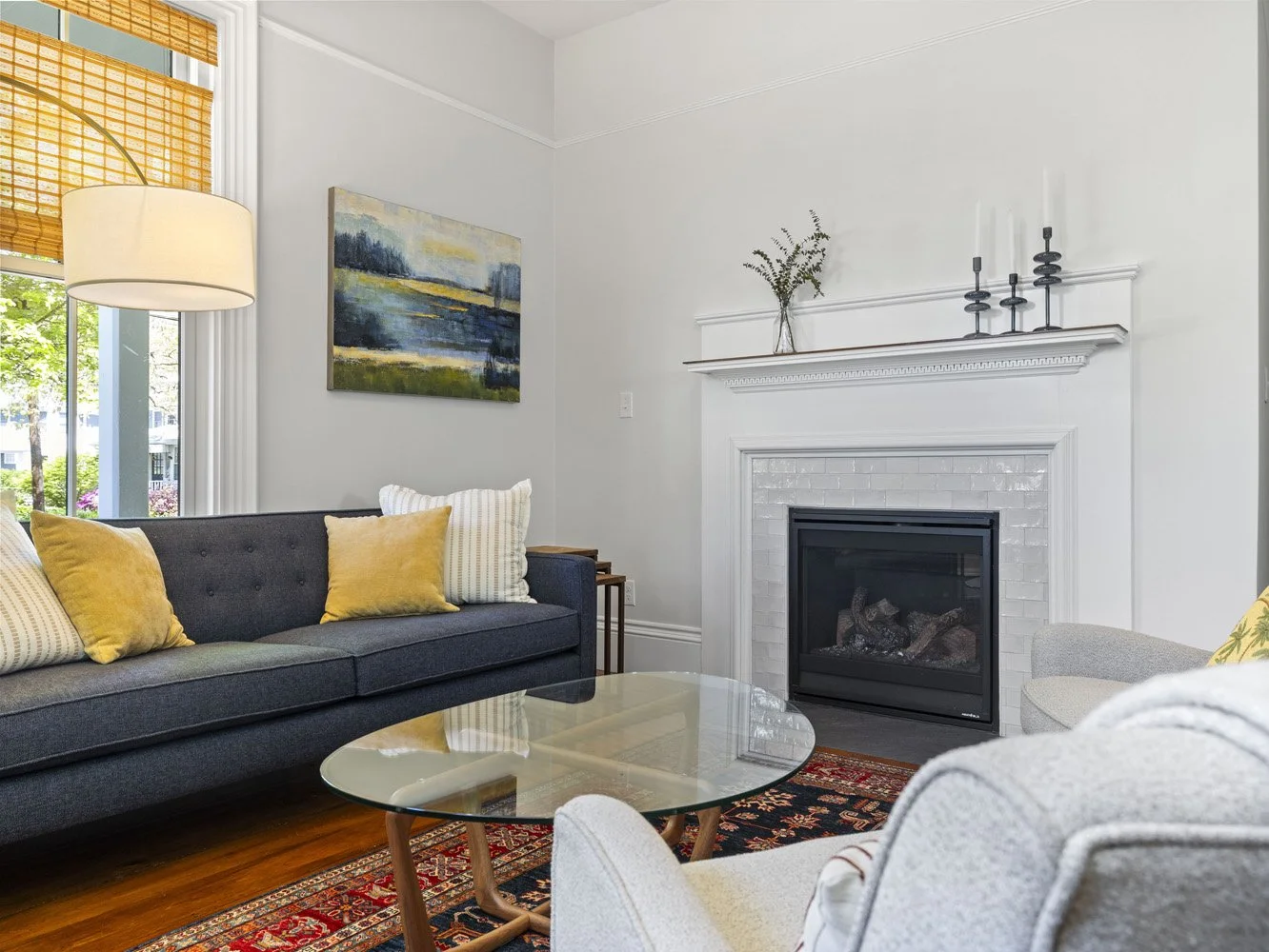 Bright living room with a dark blue sofa, mustard pillows, glass coffee table, white fireplace, and patterned rug.