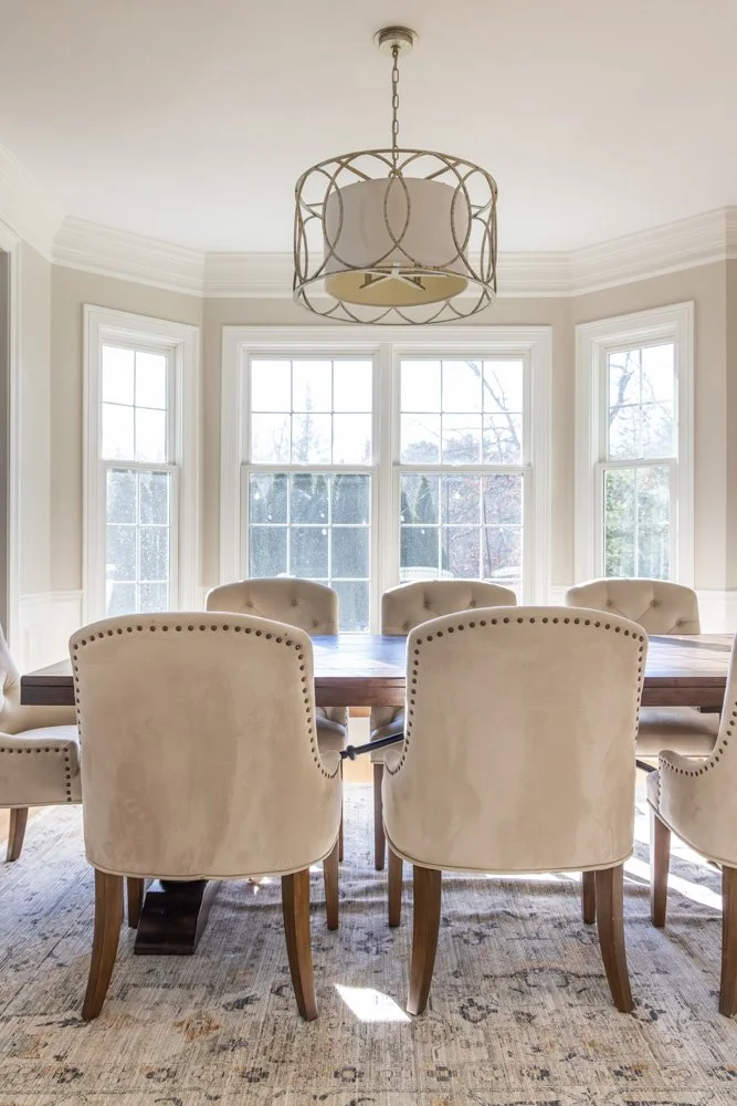 Elegant dining room with a large wooden table, upholstered nailhead chairs, bay windows, and a hanging chandelier
