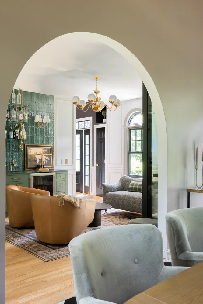 Elegant living room with arched doorway, green tile bar, chandelier, leather chairs, and gray sofa