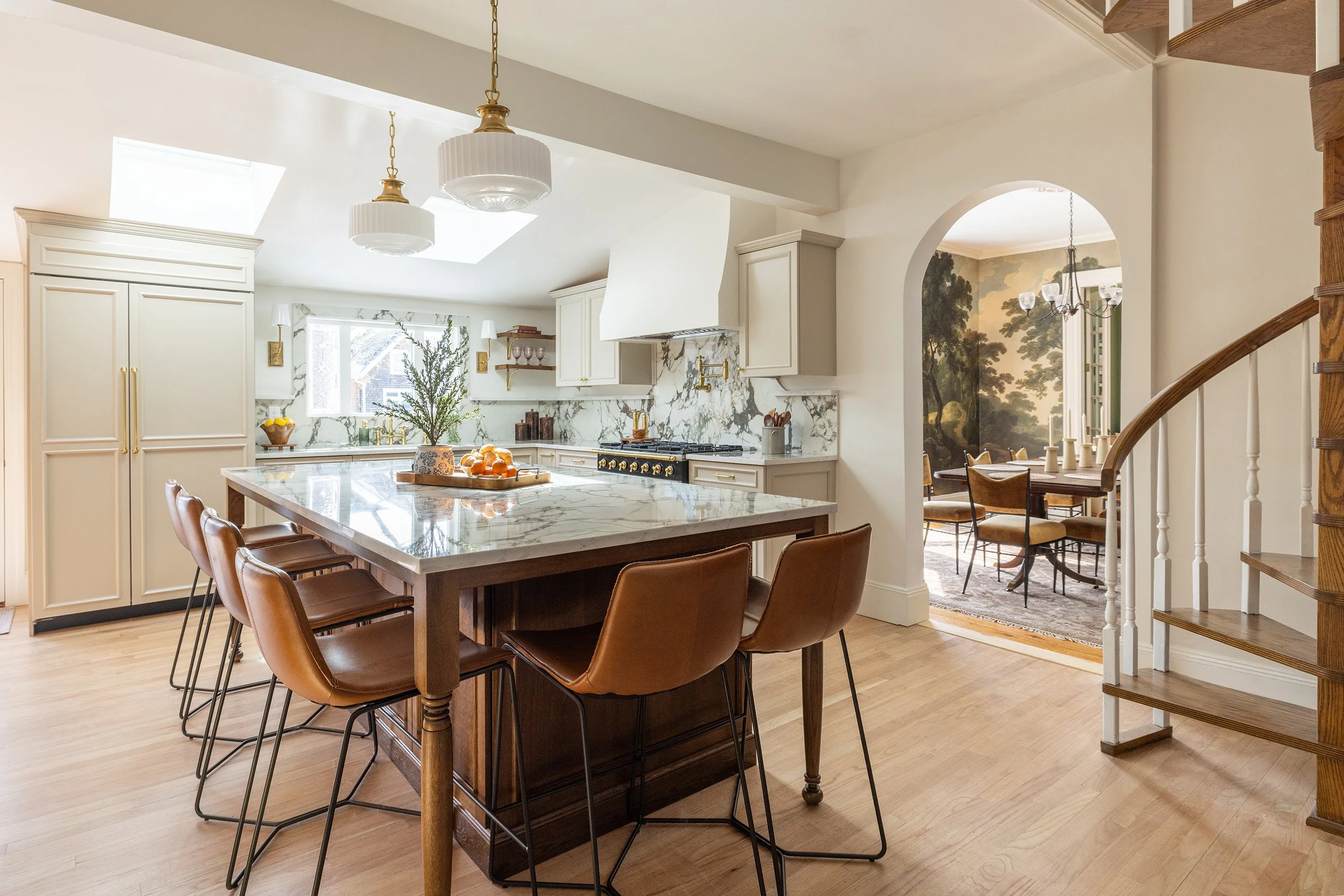Bright traditional kitchen with a marble island, leather bar stools, white cabinetry, brass fixtures, and an arched dining room beyond