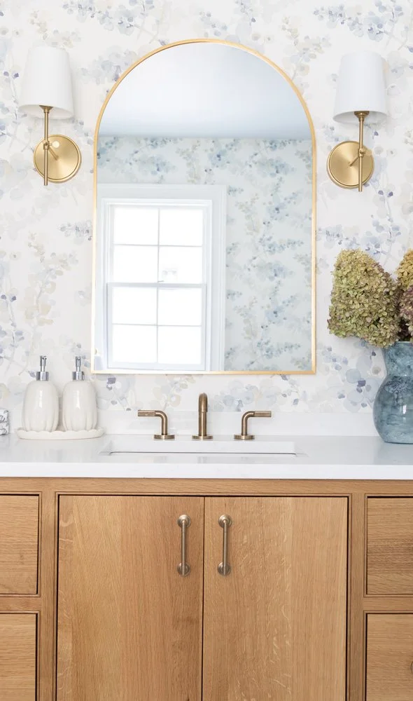 Elegant bathroom vanity with arched gold-framed mirror, floral wallpaper, brass sconces, white countertop, and wood cabinetry