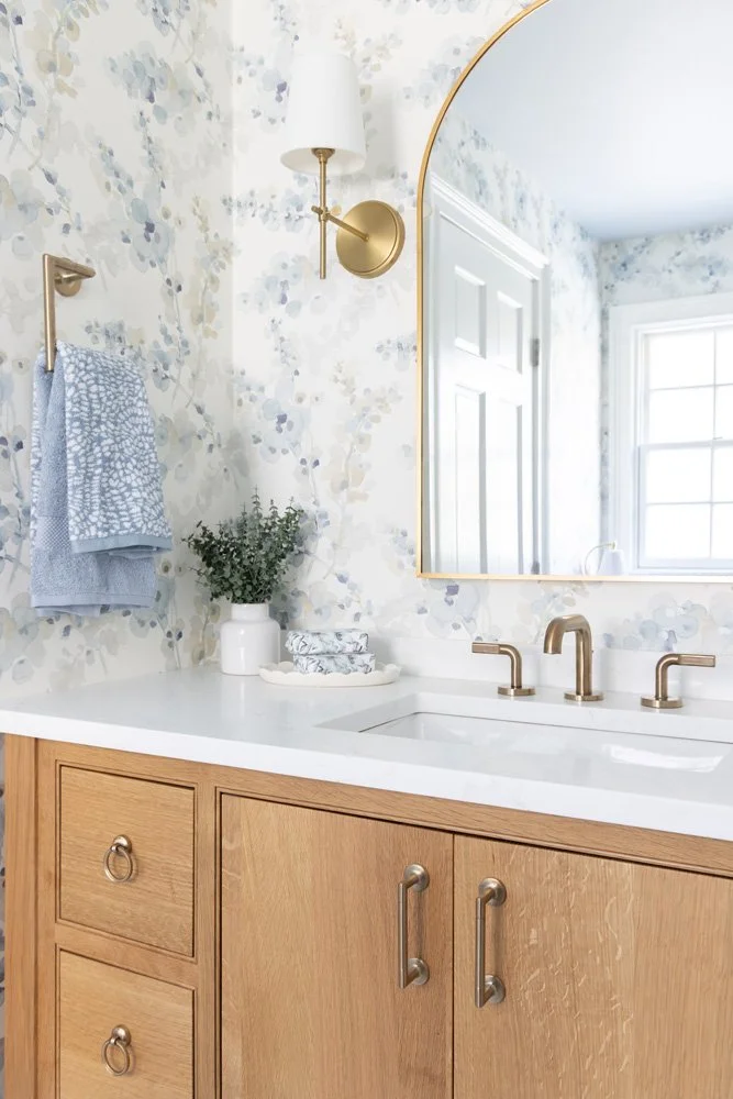 Bright bathroom vanity with white countertop, natural wood cabinets, brass fixtures, floral wallpaper, and arched mirror