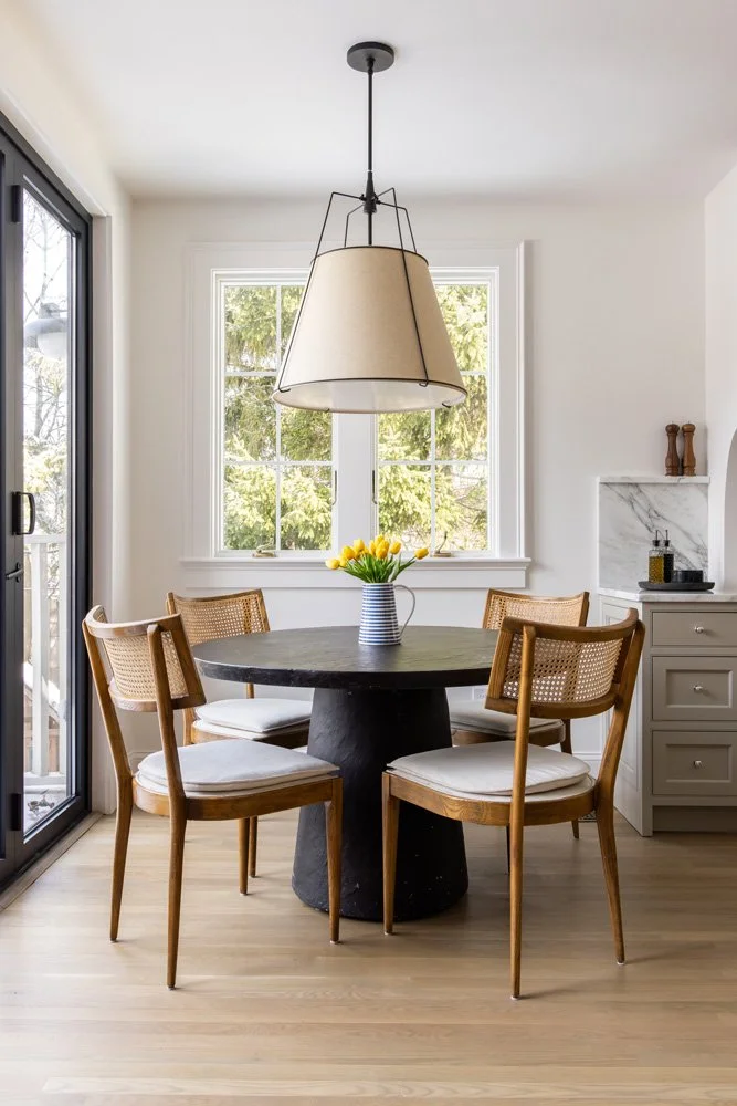 Bright dining nook with a black round table, four cane-back wooden chairs, yellow tulips, and a large pendant light by a window
