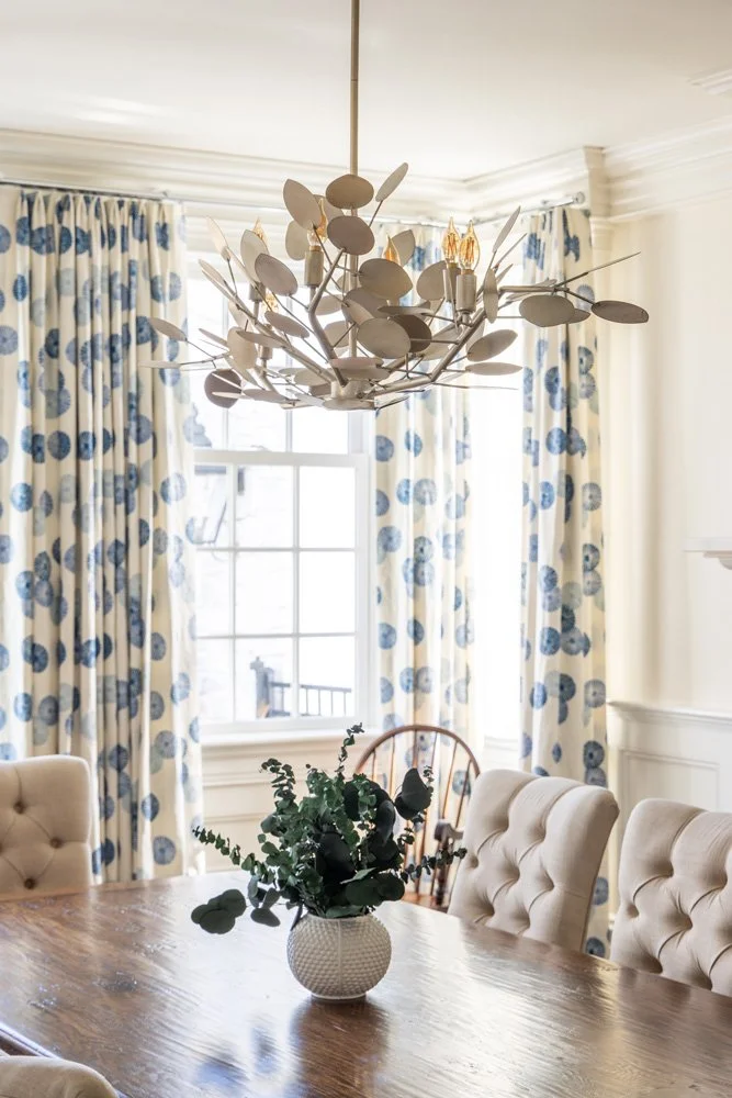 Elegant dining room with a sculptural leaf chandelier, blue-and-white patterned curtains, tufted chairs, and a vase of greenery on a wooden table